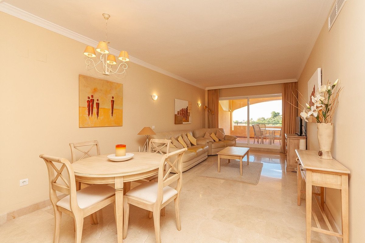 A bright living and dining area with a large sofa, dining table, and sliding doors leading to a balcony with outdoor seating.