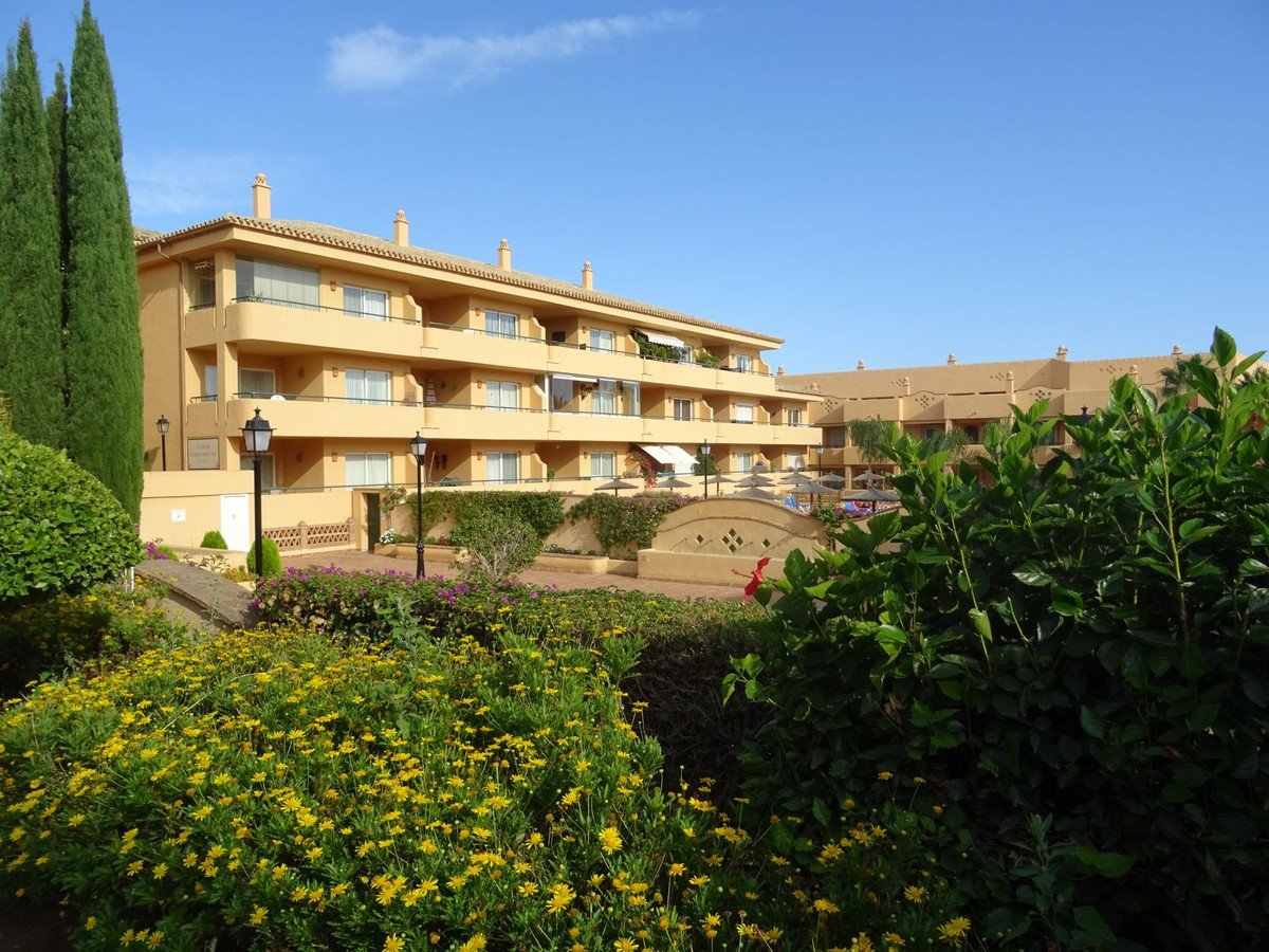 Apartment building exterior with balconies, lush gardens, and a pool area visible in the background under a clear blue sky.