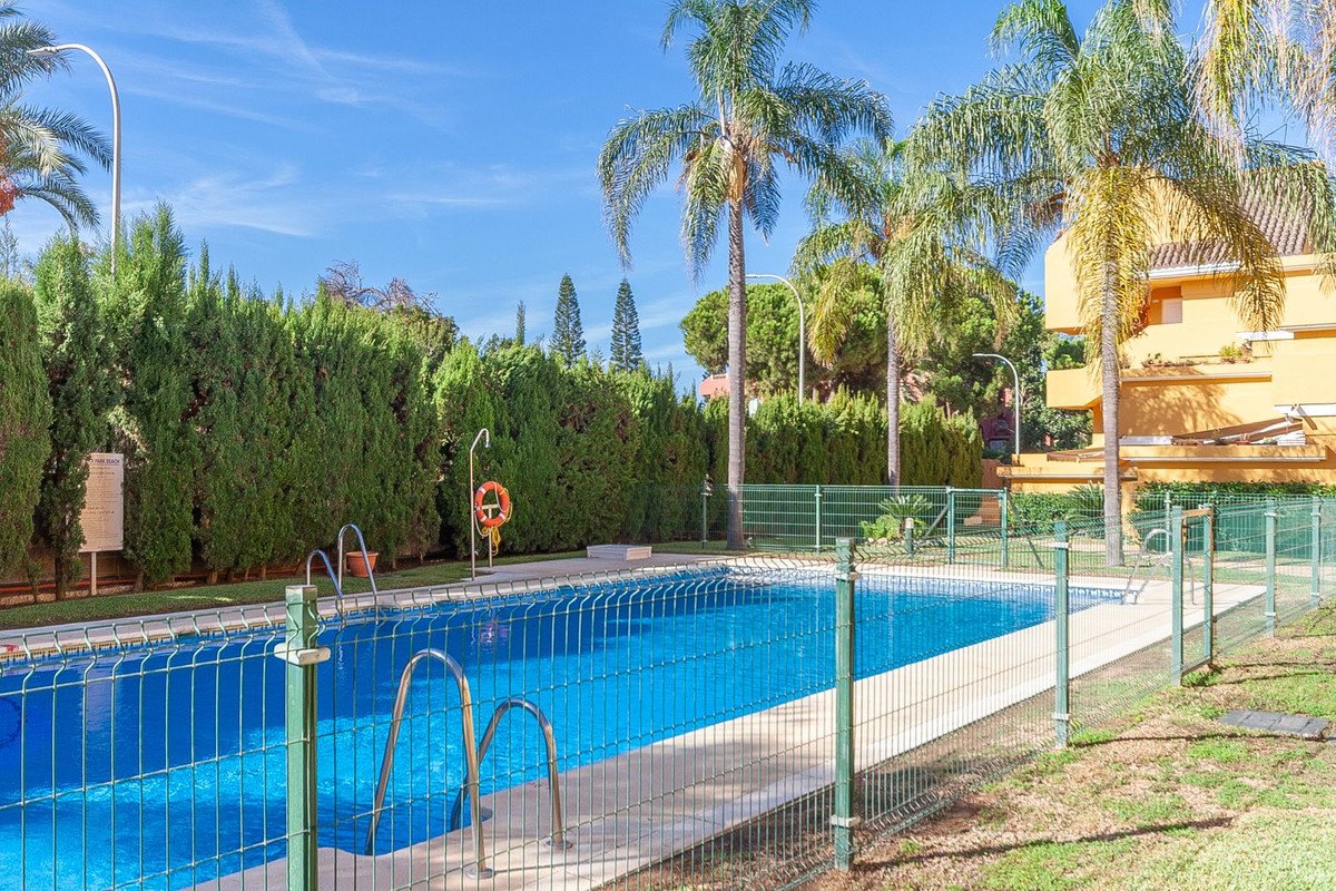 A fenced community swimming pool surrounded by lush green trees and palm trees under a clear blue sky.