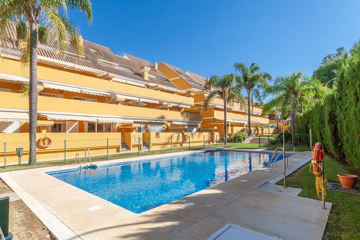 Apartment buildings with awnings and balconies overlook a bright blue swimming pool surrounded by palm trees and lush green hedges.