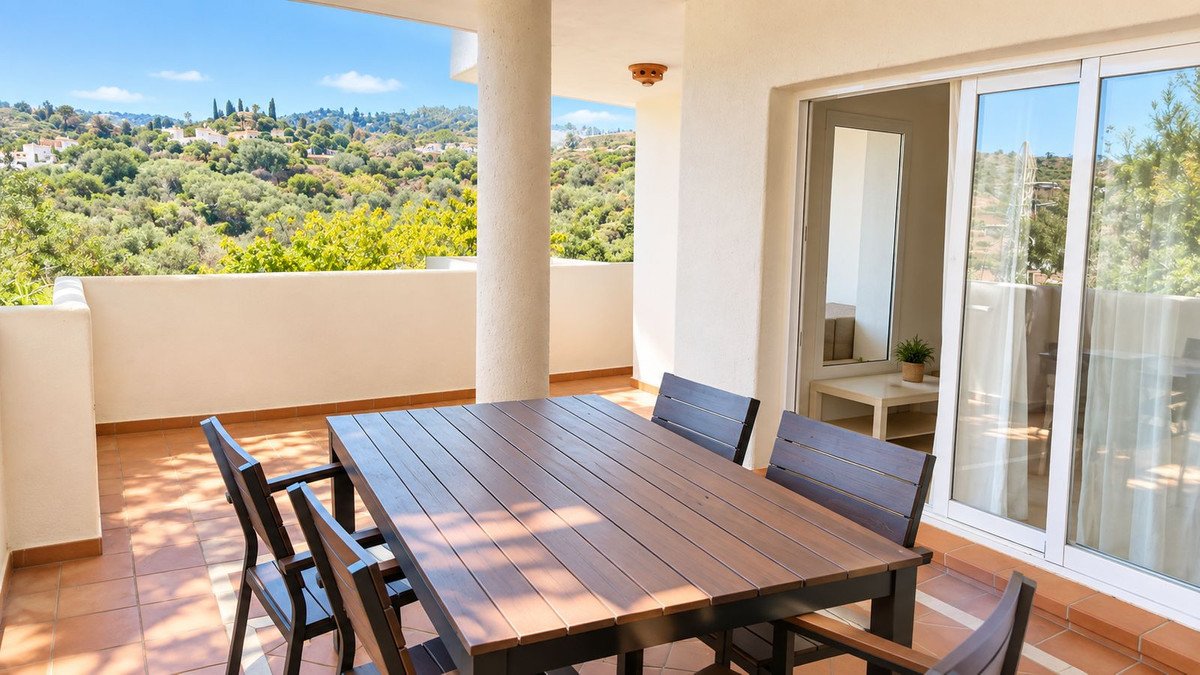 Terrace with dining table and chairs overlooking a lush, green, hilly landscape with distant white houses and a blue sky.