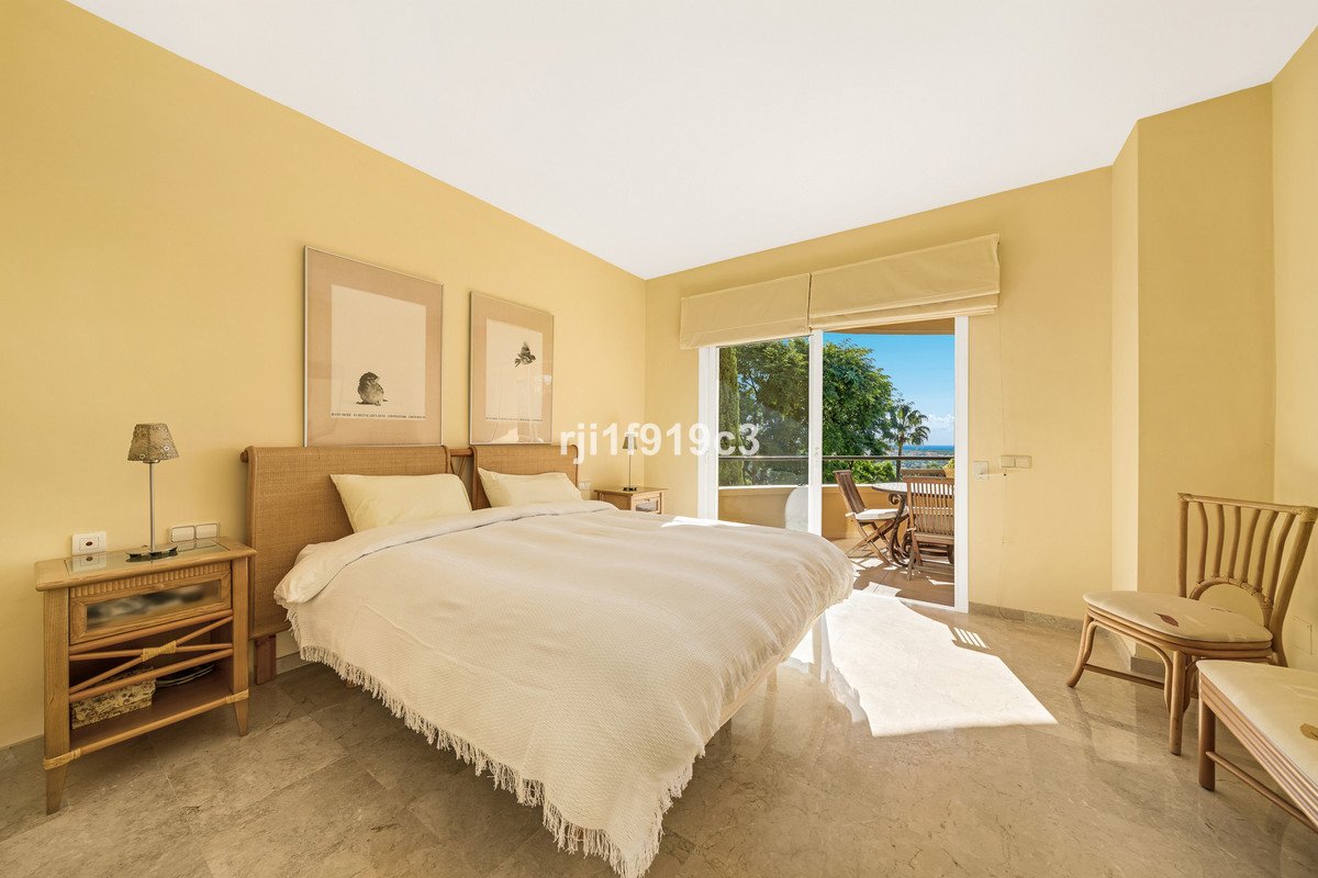 A bright bedroom with a large bed, bedside tables, and a sliding door leading to a balcony with sea view.