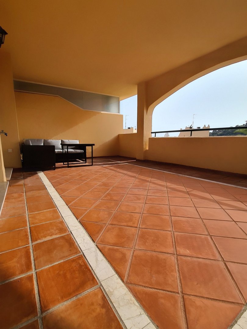 Spacious covered terrace with terracotta tiles, seating area, and a view of buildings and sky.