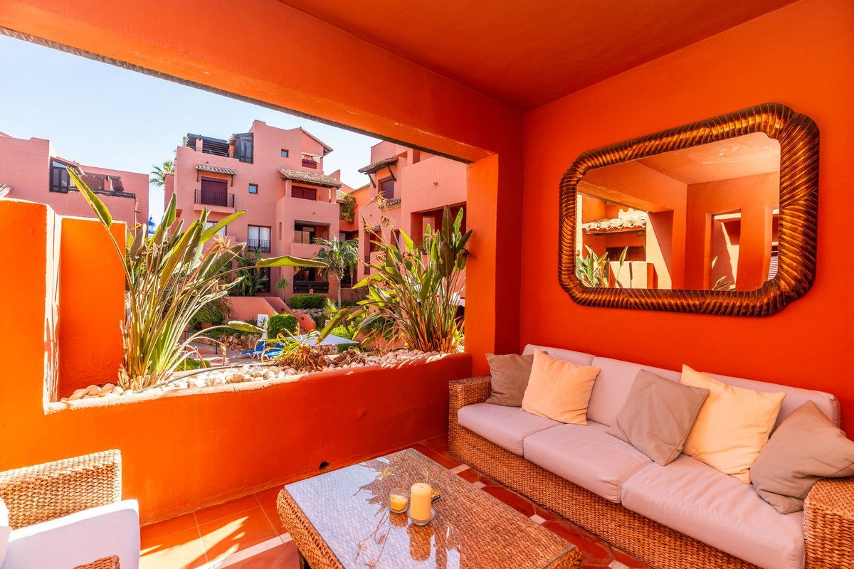 Terrace with a sofa and coffee table, overlooking a courtyard with tropical plants and buildings.