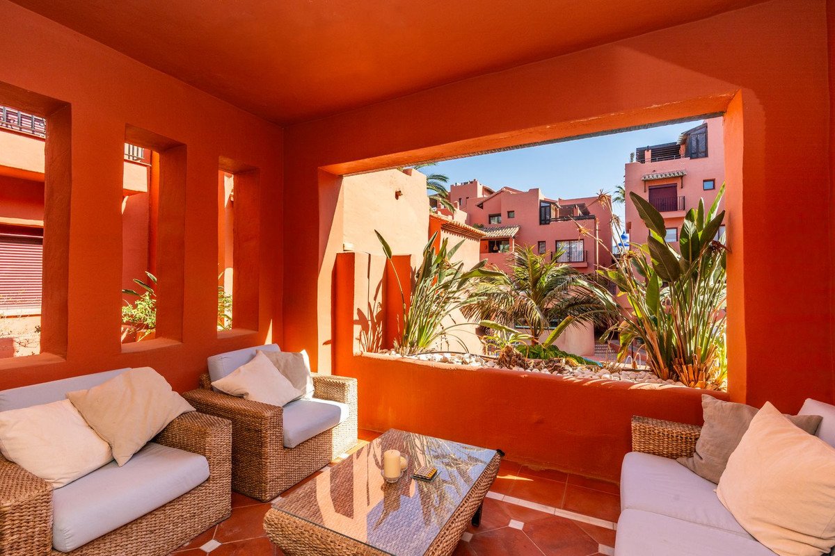 Orange terrace with wicker sofas and a glass coffee table, overlooking a courtyard with palm trees and apartments.
