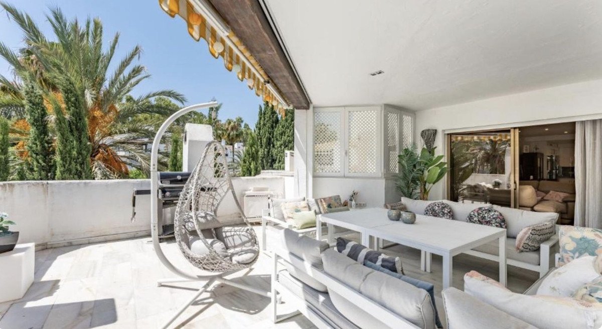 Spacious white terrace with outdoor dining, lounge seating, hanging chair, and a grill, overlooking palm trees.