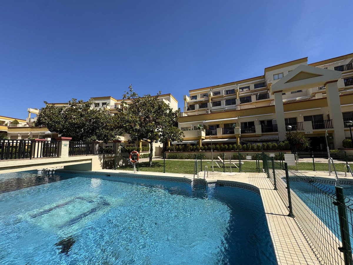 A sparkling blue swimming pool in front of a multi-story apartment complex with balconies and trees under a clear blue sky.