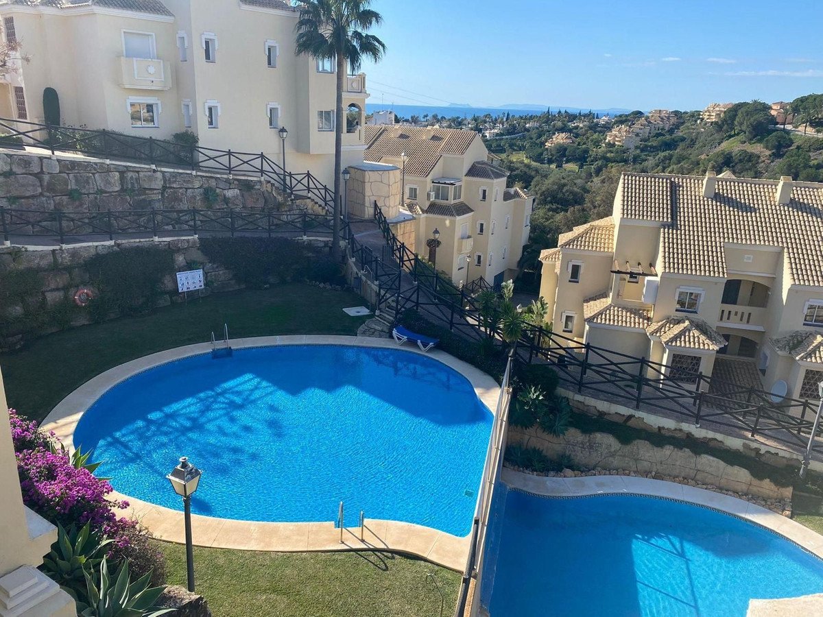 Two blue swimming pools with surrounding green grass and apartments, overlooking the sea in the distance on a sunny day.