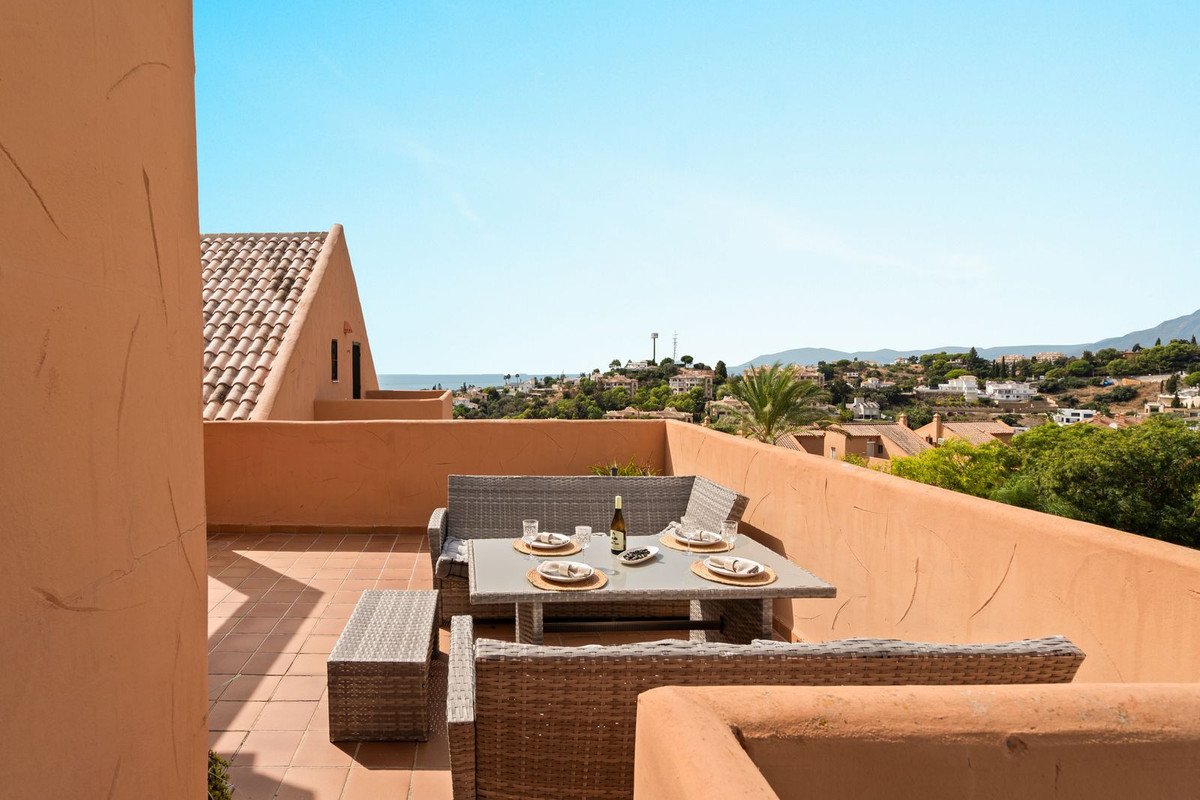 Rooftop terrace with dining set and sofa, overlooking a town with distant sea and mountains under a clear blue sky.