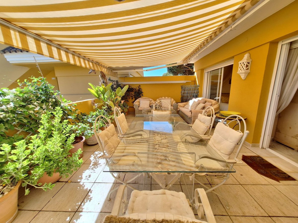 Sunlit patio with glass table, white chairs, yellow striped awning, and potted plants.