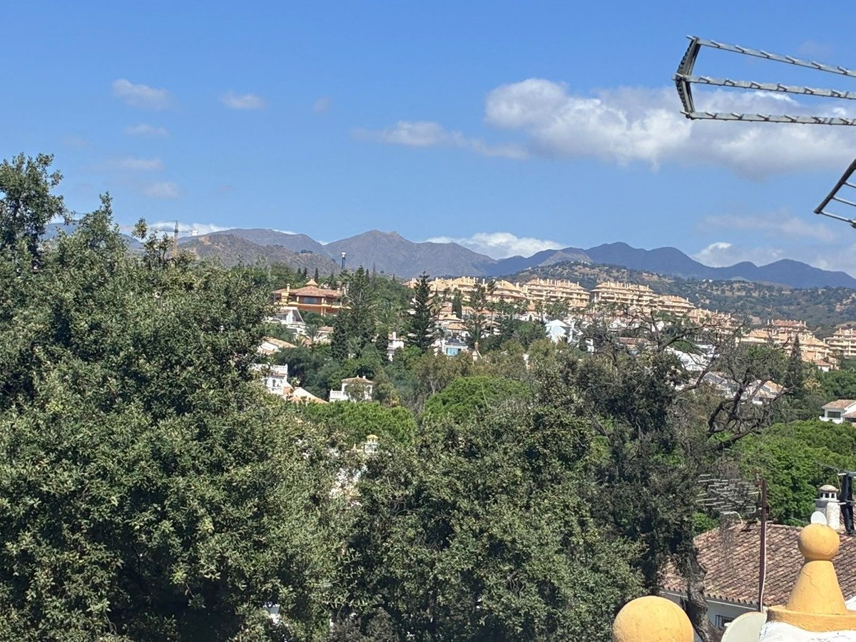 Scenic view of a village nestled in hills with mountains in the background, surrounded by lush green trees under a blue sky.