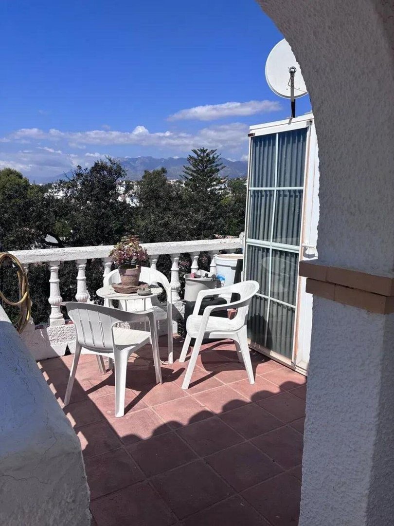 A sunny balcony with white plastic chairs and table, overlooking trees and distant mountains.