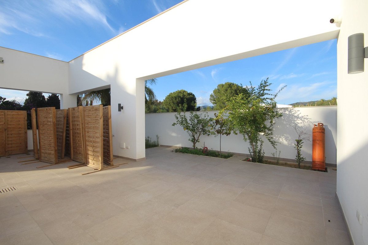Modern patio with a small garden, trees, and a clear blue sky with clouds. Gas cylinder and fence panels are visible.