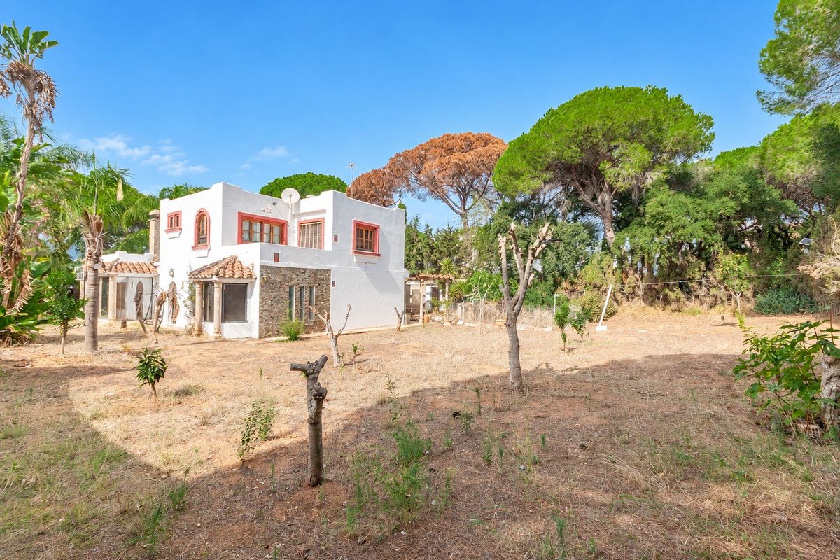 A white two-story villa with red trim, surrounded by tropical trees and dry grass under a clear blue sky.