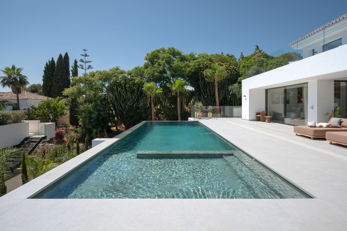 Modern villa with an infinity pool and sun loungers on a spacious terrace with lush green gardens and blue sky.