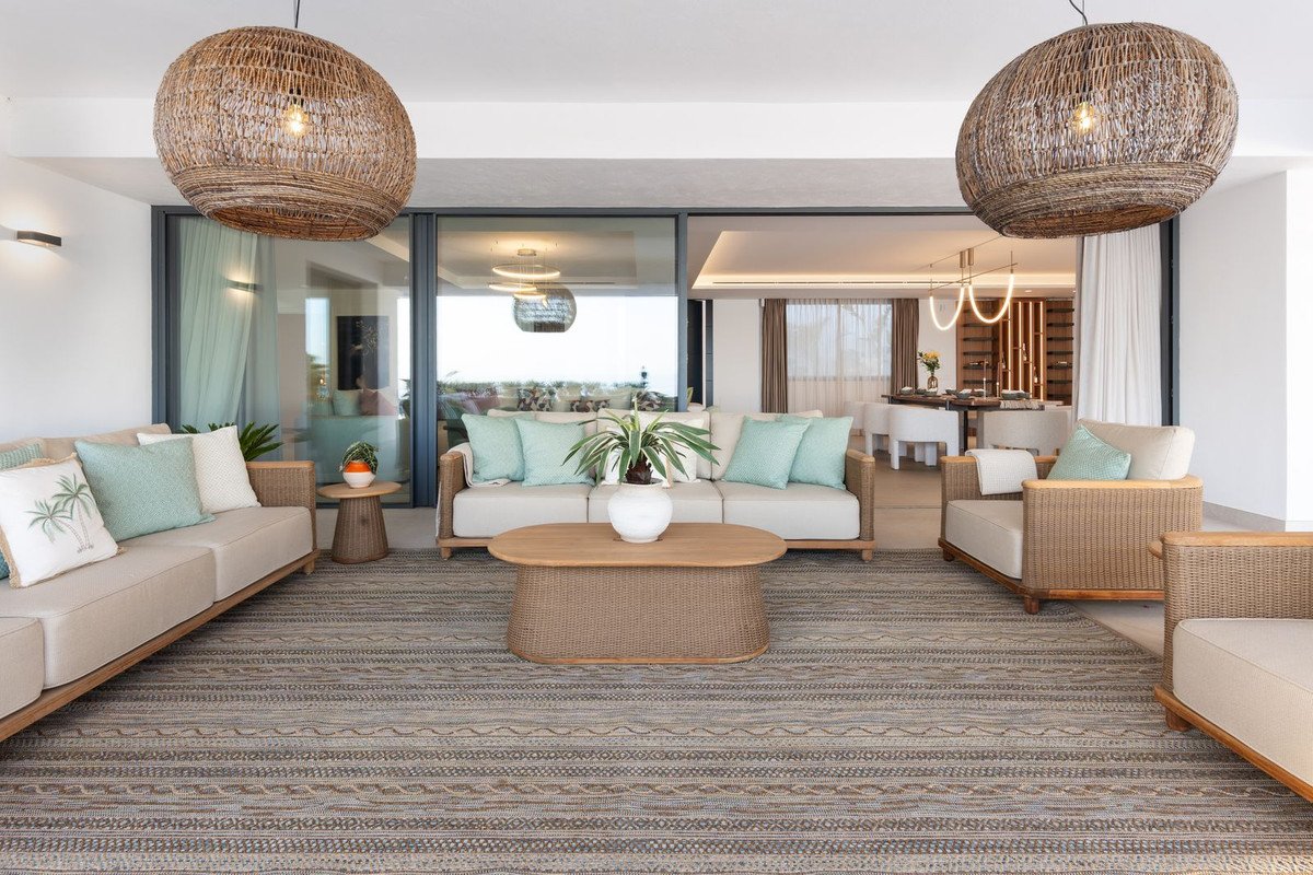 Spacious covered patio with rattan furniture, light-colored sofas, and a coffee table, overlooking a bright living and dining area with ocean views.