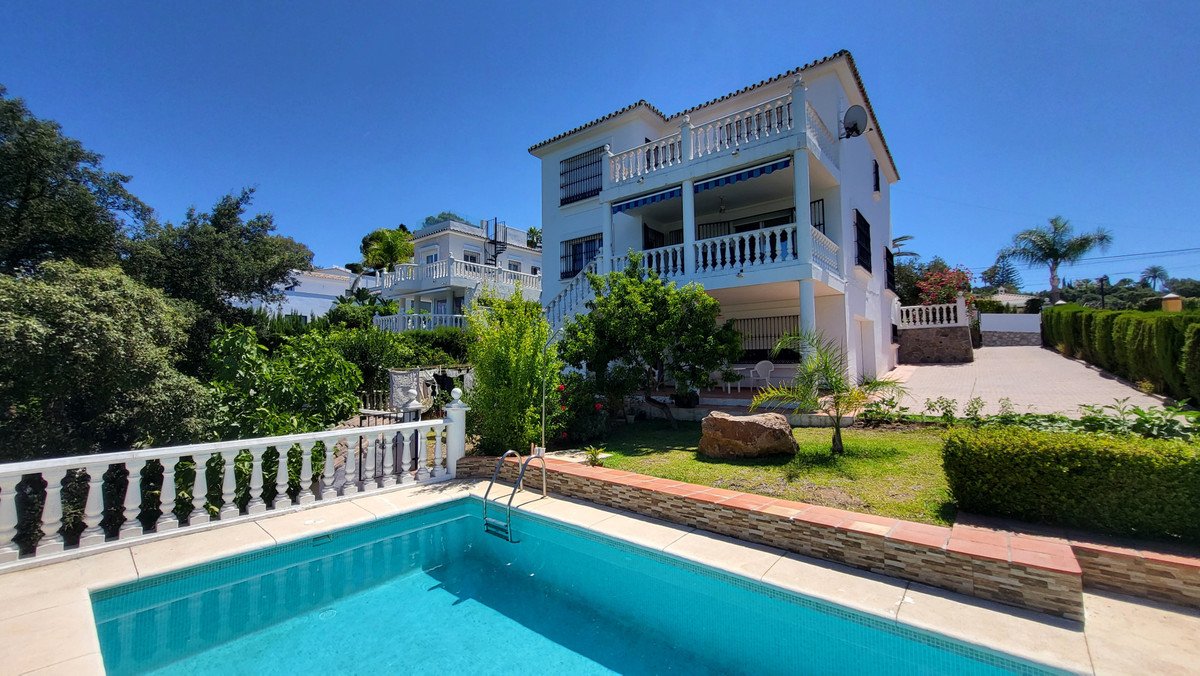 A multi-story white villa with balconies and awnings, overlooking a blue swimming pool and a lush green garden under a clear blue sky.