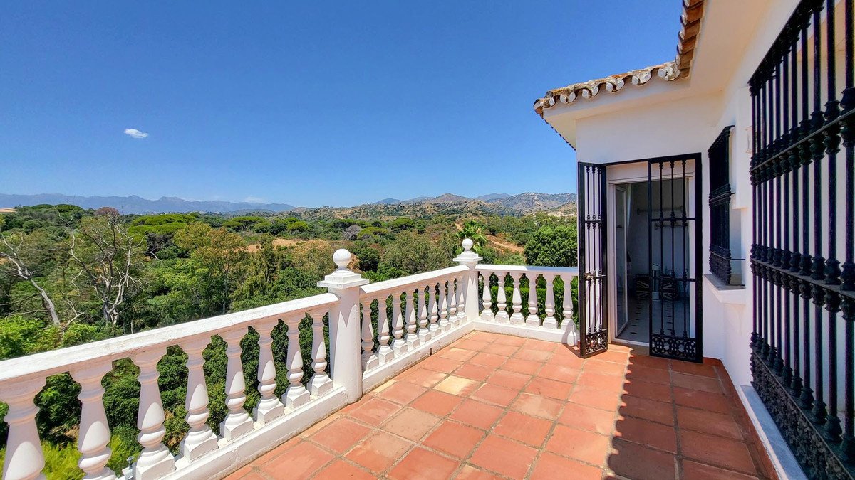 A white balustrade railing on a terracotta tiled terrace overlooking lush green trees and distant mountains under a clear blue sky.