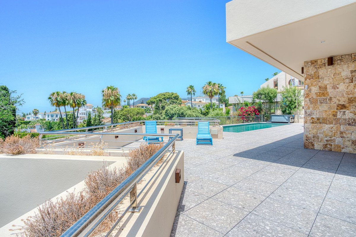 Modern terrace with sun loungers, swimming pool, and a distant view of palm trees and houses under a clear blue sky.