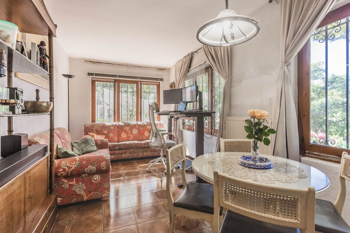 Living and dining area with patterned sofas, a desk with dual monitors, and a round table with chairs, featuring a garden view through large windows.