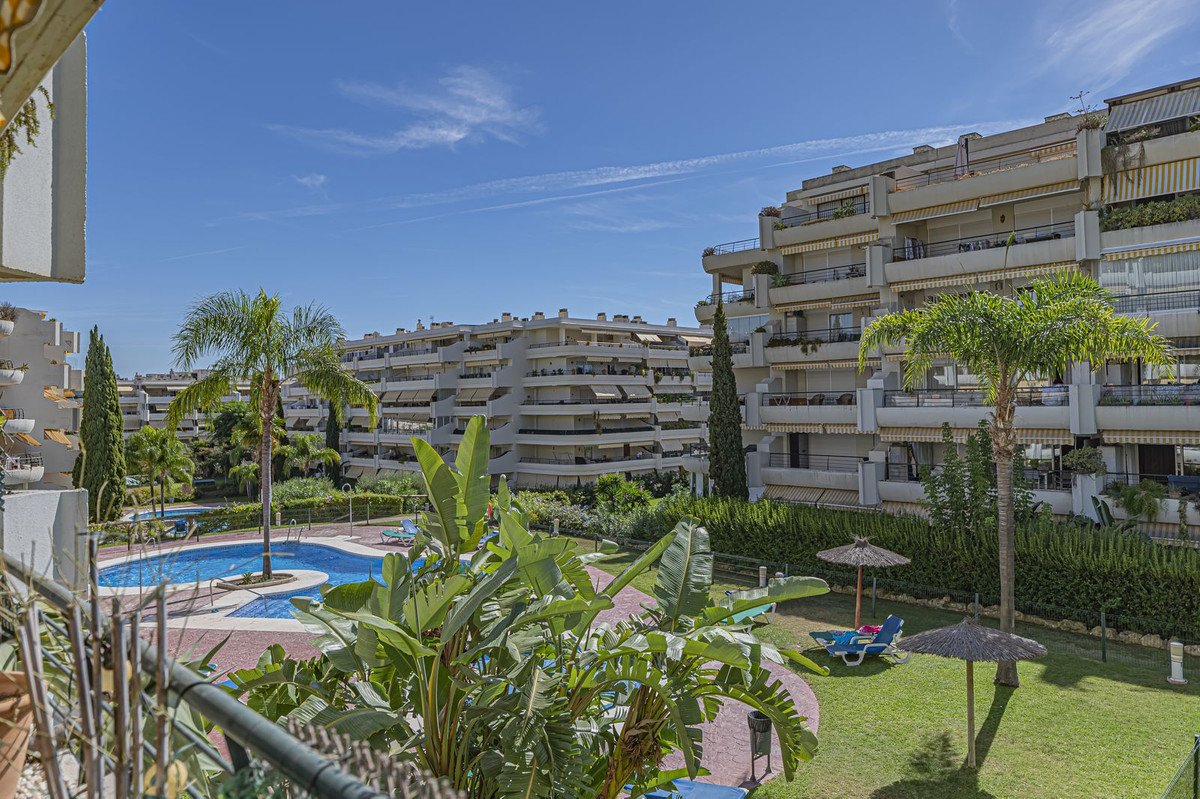 View of a residential complex with a swimming pool, sun loungers, palm trees, and lush greenery under a blue sky.