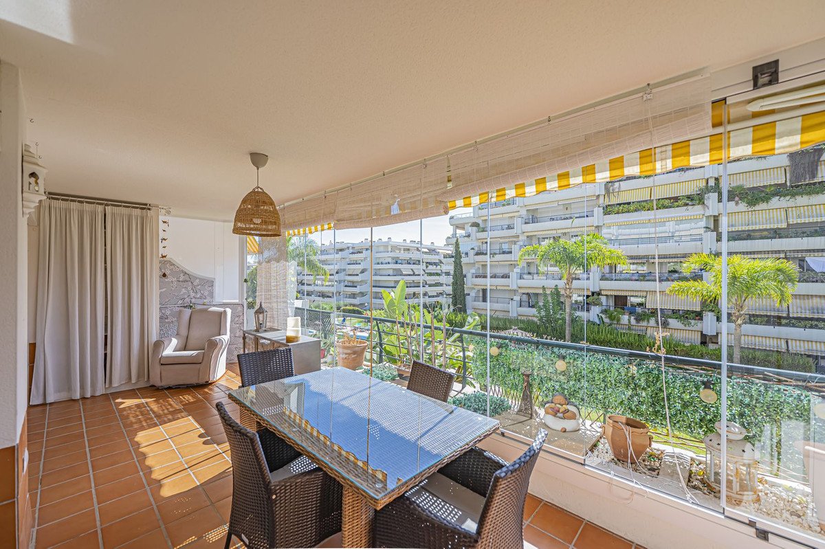 Enclosed terrace with dining table and chairs, offering a view of apartment buildings and greenery.