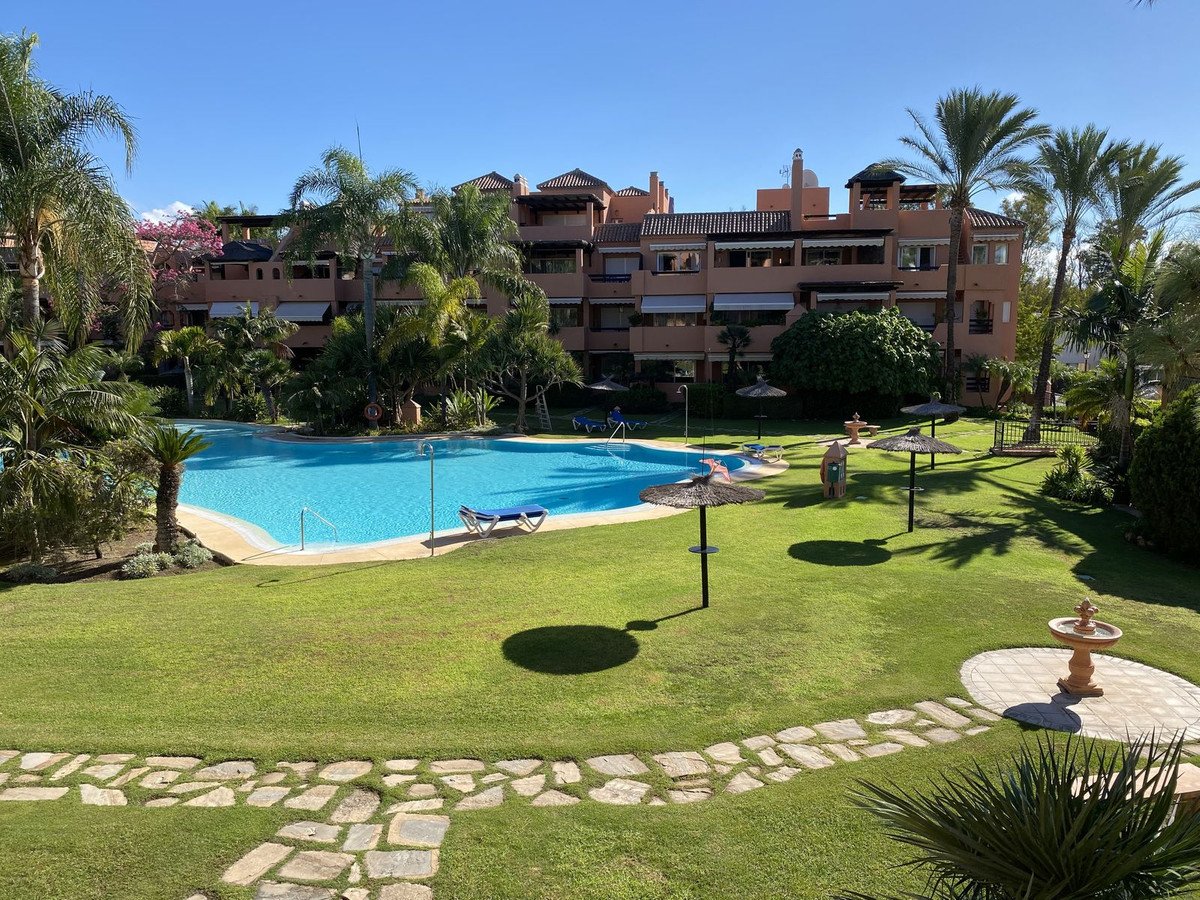 A large swimming pool and green lawn with sun loungers and umbrellas, in front of a multi-story apartment building.