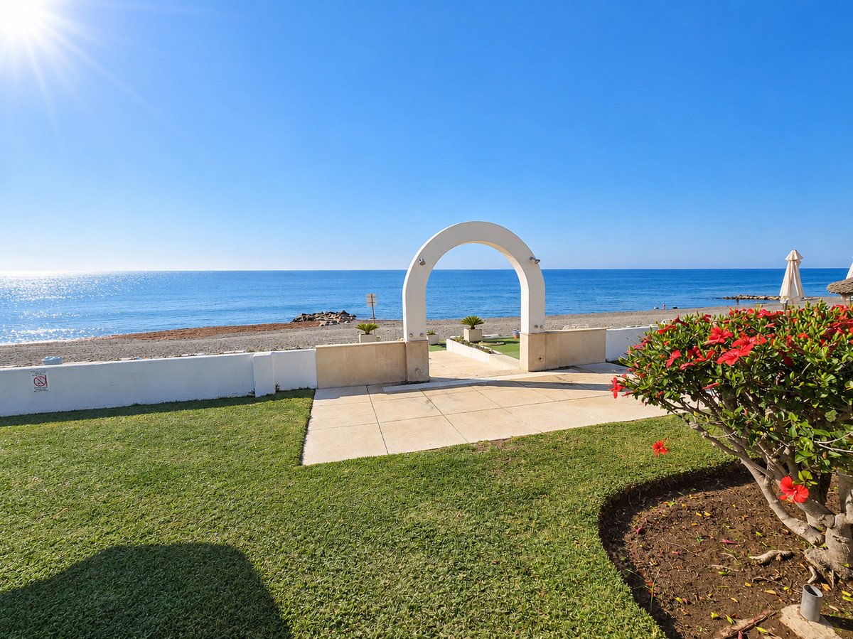 A beautiful garden area with green grass and flowering bushes leads to an archway with a stunning view of the blue ocean and beach.