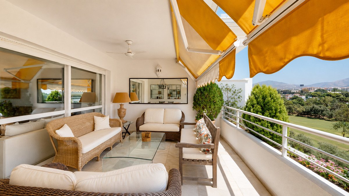 Spacious terrace with rattan seating, glass coffee table, and sunny yellow awnings, overlooking a green golf course and distant hills.