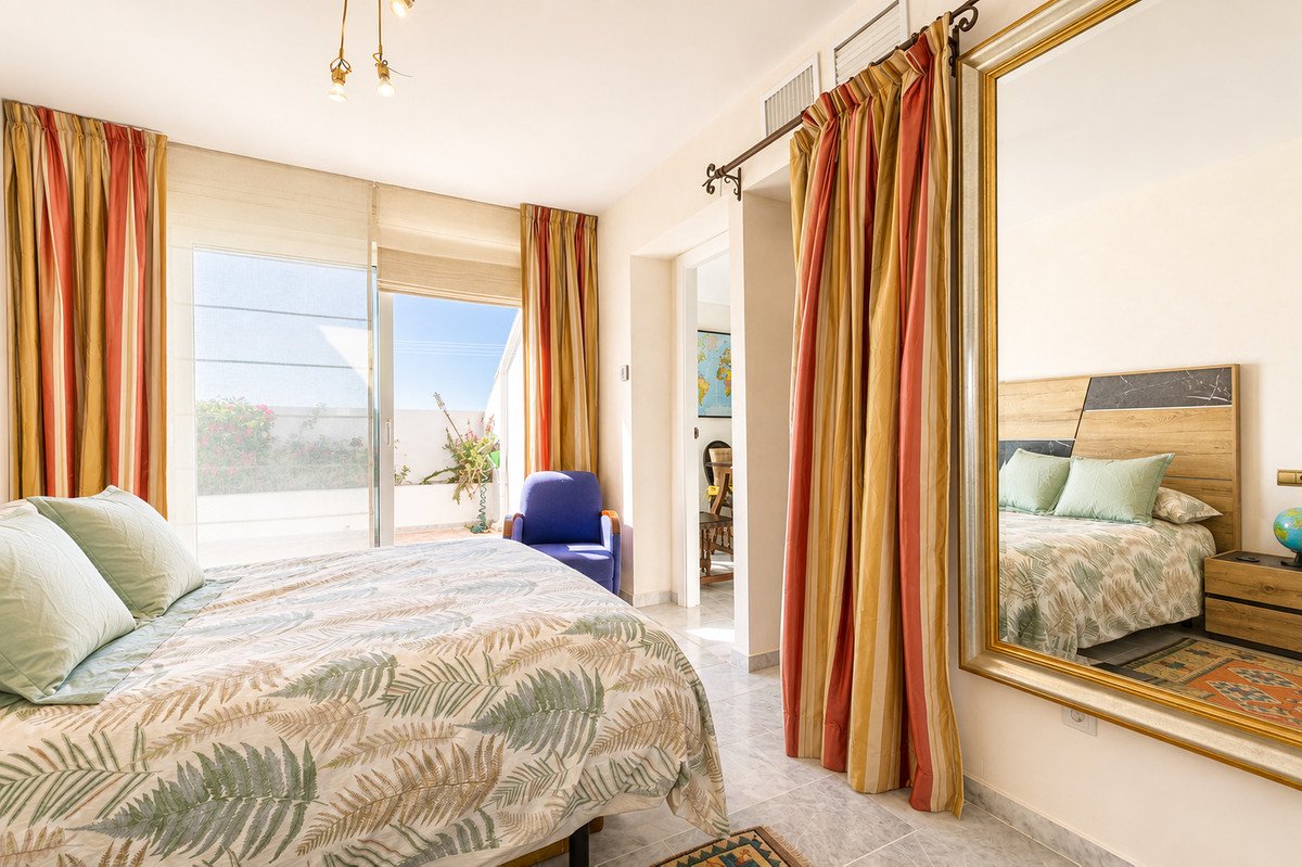 A bright bedroom with a king-size bed, tropical bedding, striped curtains, and a glass door leading to a terrace with a blue armchair.