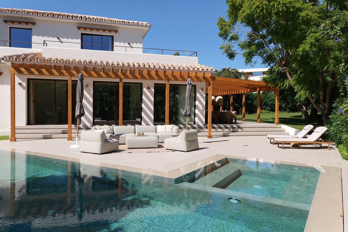 Luxurious modern villa with a sparkling pool, outdoor lounge area under a wooden pergola, and sun loungers on a sunny day.