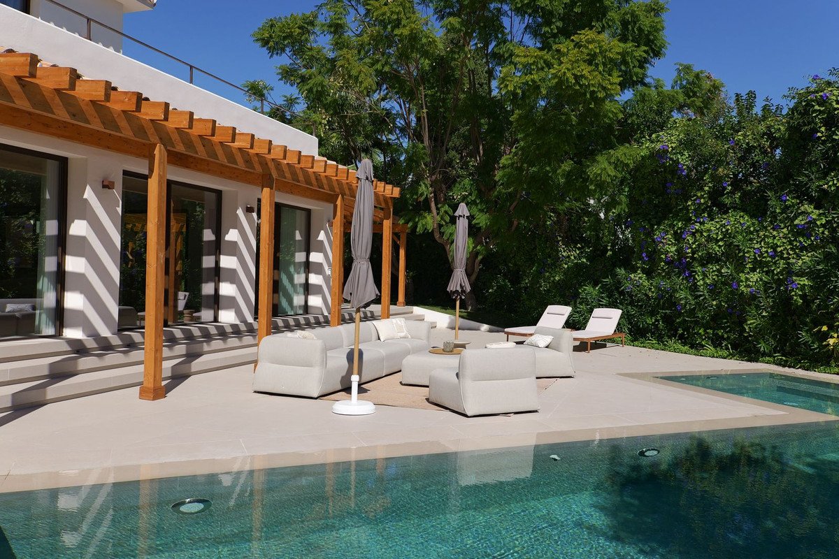 Outdoor lounge seating and pool area with a wooden pergola and lush greenery under a clear blue sky.