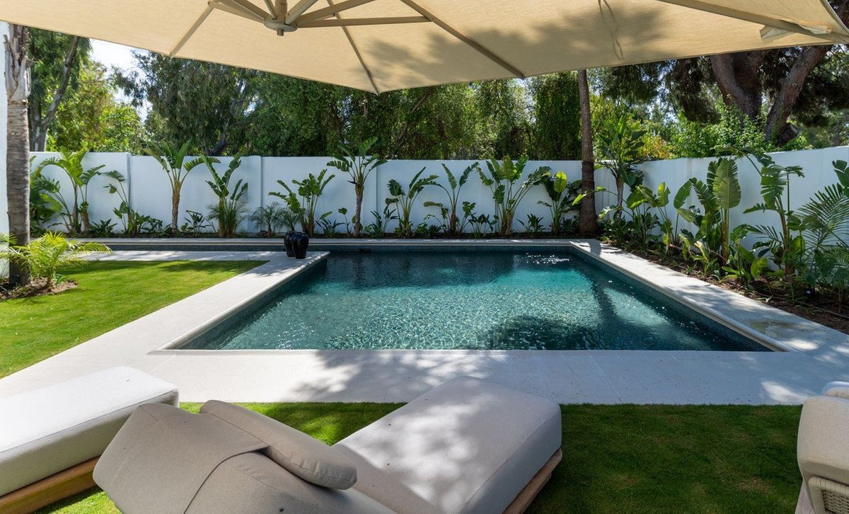 Backyard pool with lounge chairs, green lawn, and tropical plants under a large umbrella