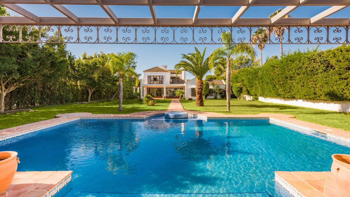 Luxury villa with a large swimming pool, lush green garden, palm trees, and a clear blue sky.