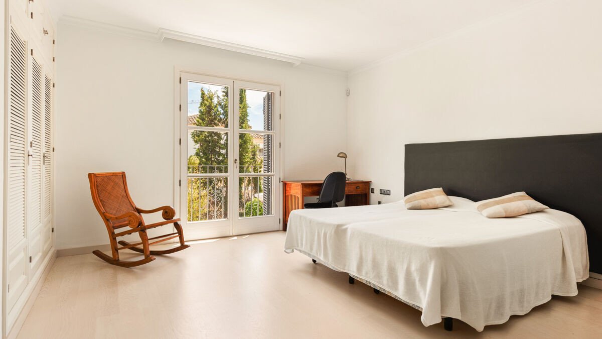A bright bedroom with two beds, a rocking chair, a desk with a chair, and a balcony door with a green view.