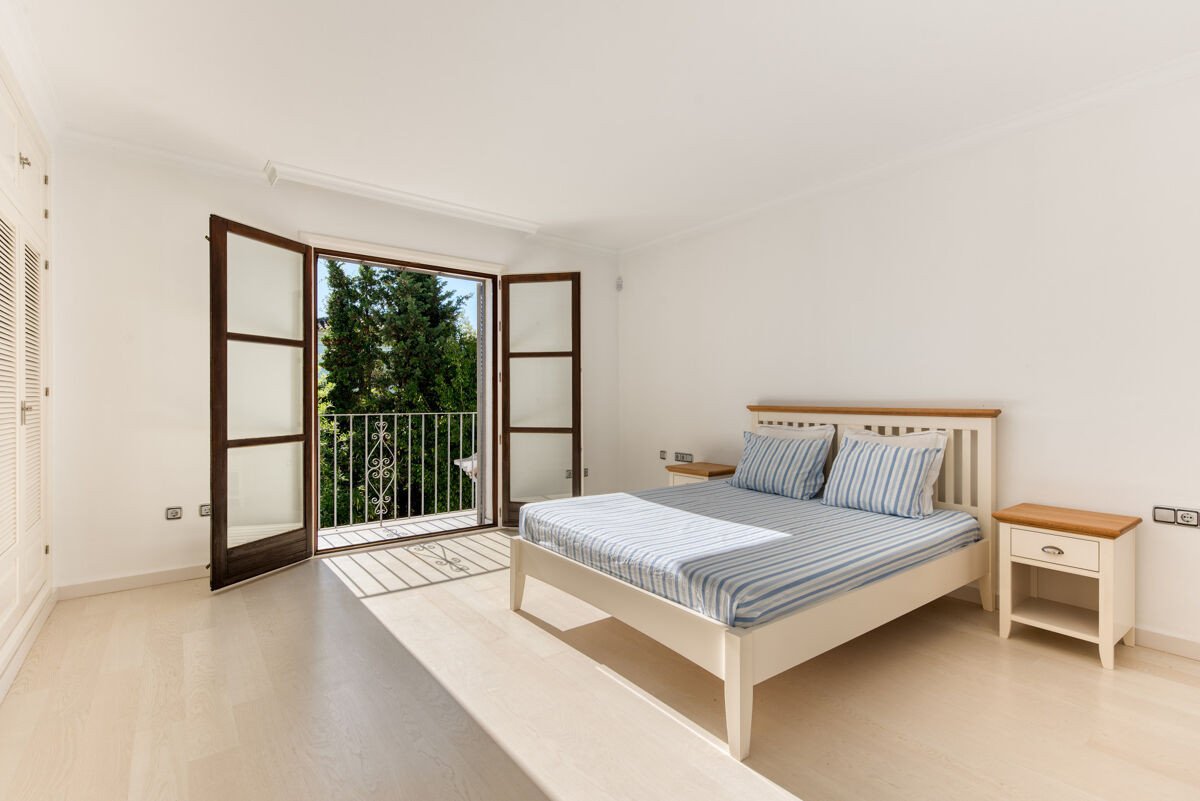 A spacious bedroom with a double bed featuring blue and white striped bedding, opening to a balcony with a green tree view.