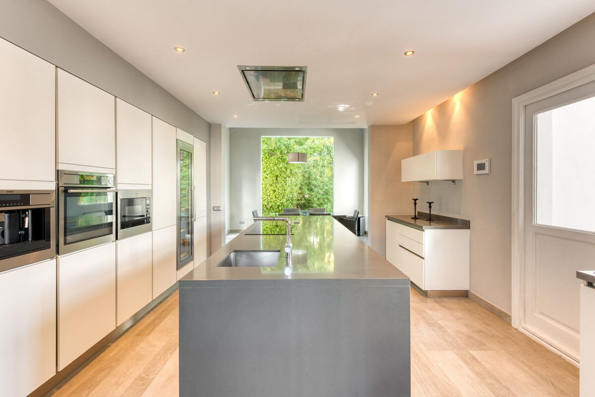 A modern, spacious kitchen featuring a large island with a sink, integrated appliances, and a view of lush greenery through a big window.