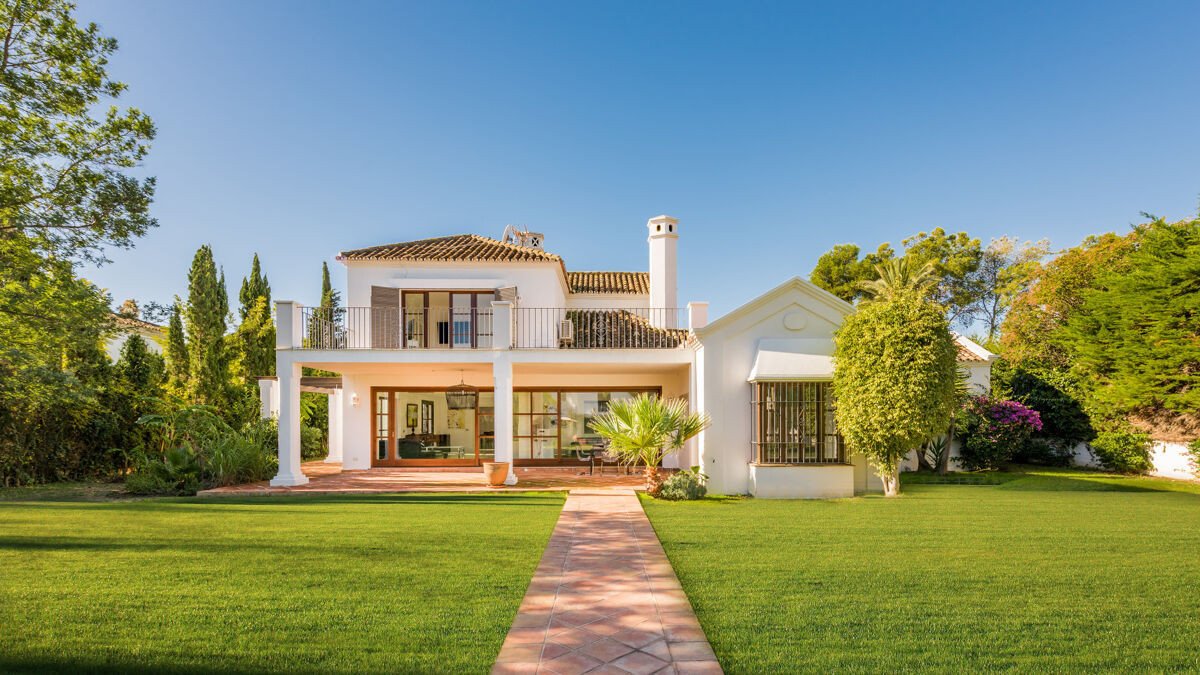 A white villa with a balcony, large windows, and a lush green lawn leading to the entrance.