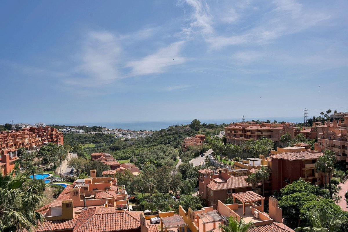View from a balcony overlooking a complex with terracotta roofs, lush greenery, a swimming pool, and the sea in the distance.