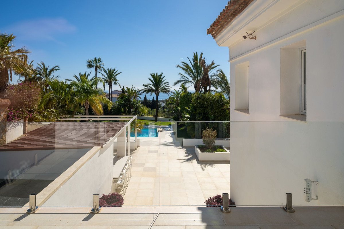 Modern terrace with glass railings overlooking a pool, lush palm trees, and a distant sea view.