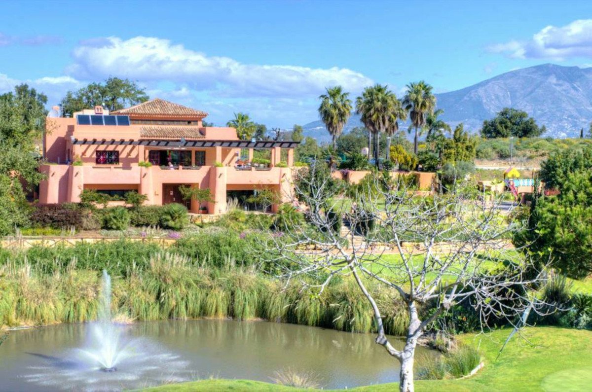 A large coral villa with a balcony, overlooking a pond with a fountain and lush green gardens with mountains in the distance.