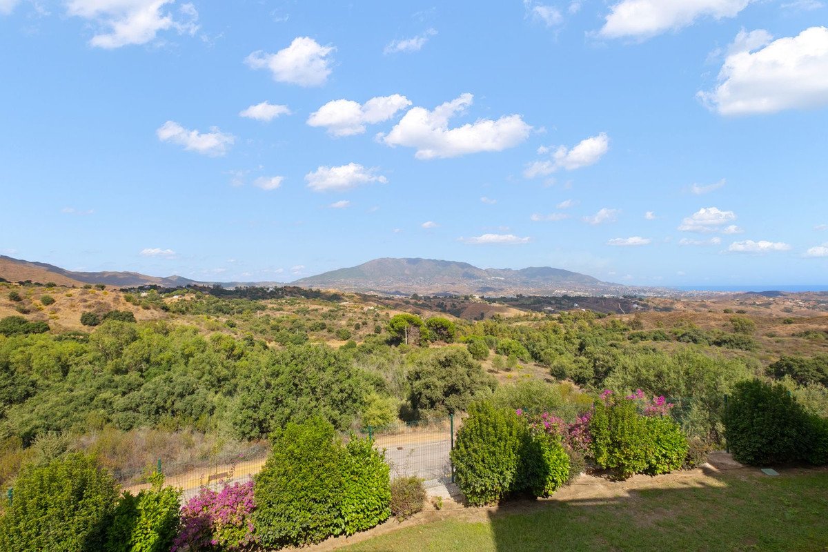 Expansive view of rolling hills, lush greenery, distant mountains, and the sea under a blue sky with white clouds.