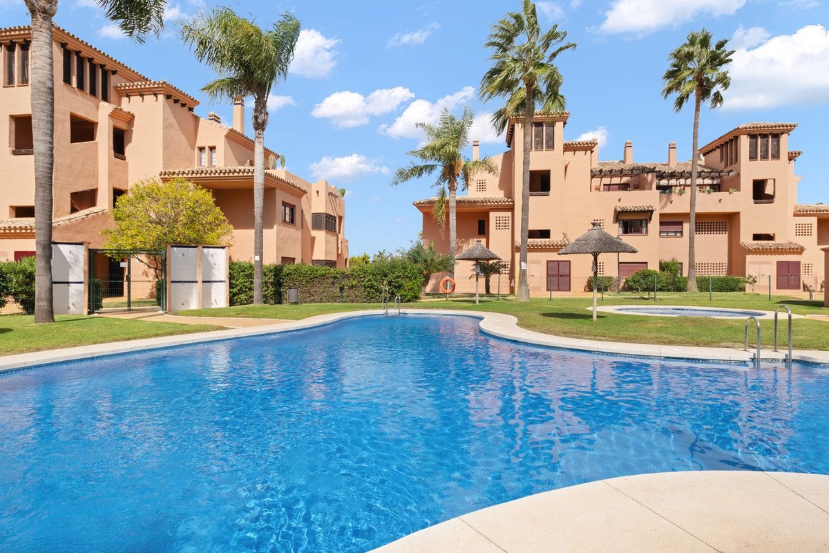 Resort buildings with palm trees surround a large, inviting blue swimming pool on a sunny day.