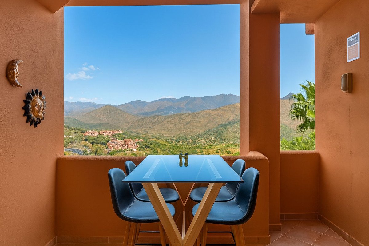 Balcony with glass table and blue chairs, overlooking mountainous landscape and village under clear blue sky.