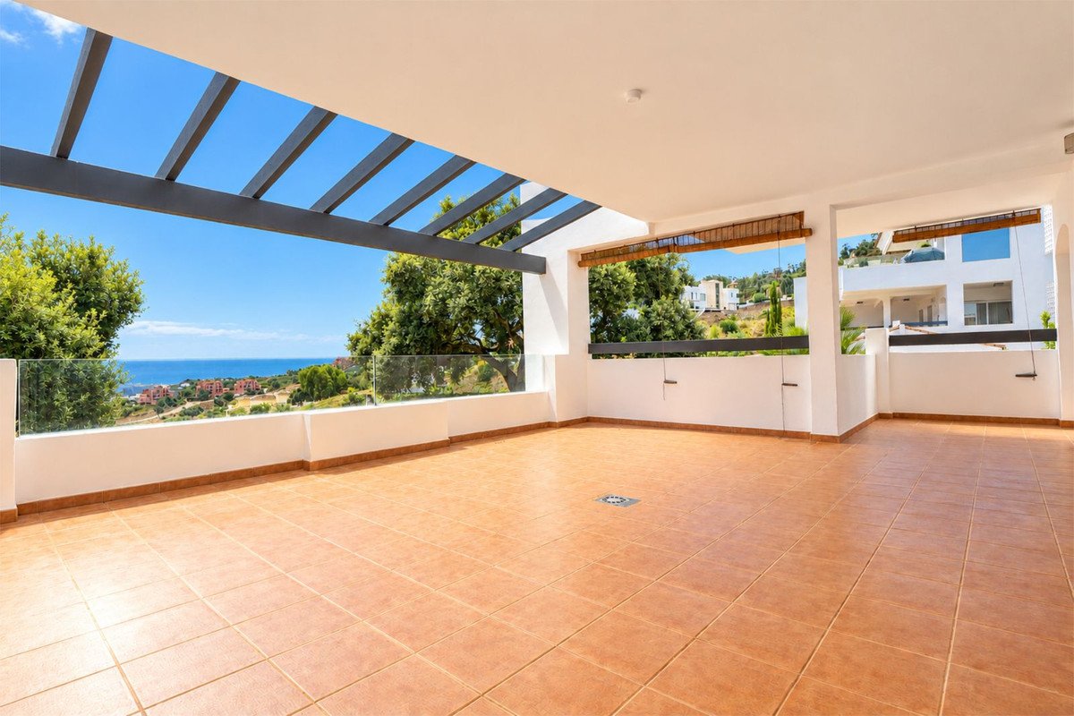 Spacious tiled terrace with a pergola, overlooking the sea and greenery, with a modern building in the background.