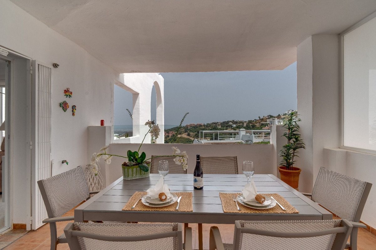 Terrace dining table set for two with a view of hills and distant sea, featuring potted plants and white architecture.