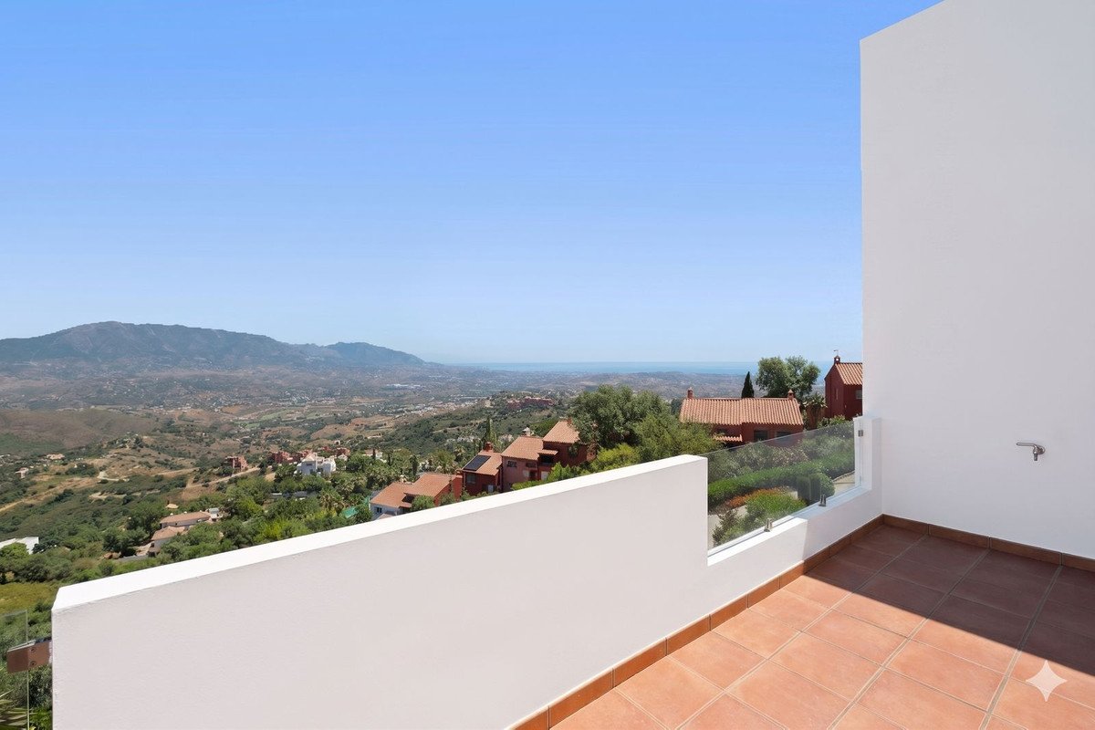 A modern terrace with a white wall and terracotta tiles offers a stunning panoramic view of mountains, greenery, and the distant sea under a clear blue sky.
