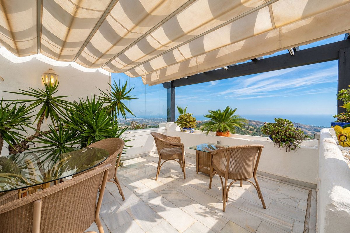 Terrace with wicker seating and tables, overlooking the sea and distant town, with lush green plants and a retractable awning.