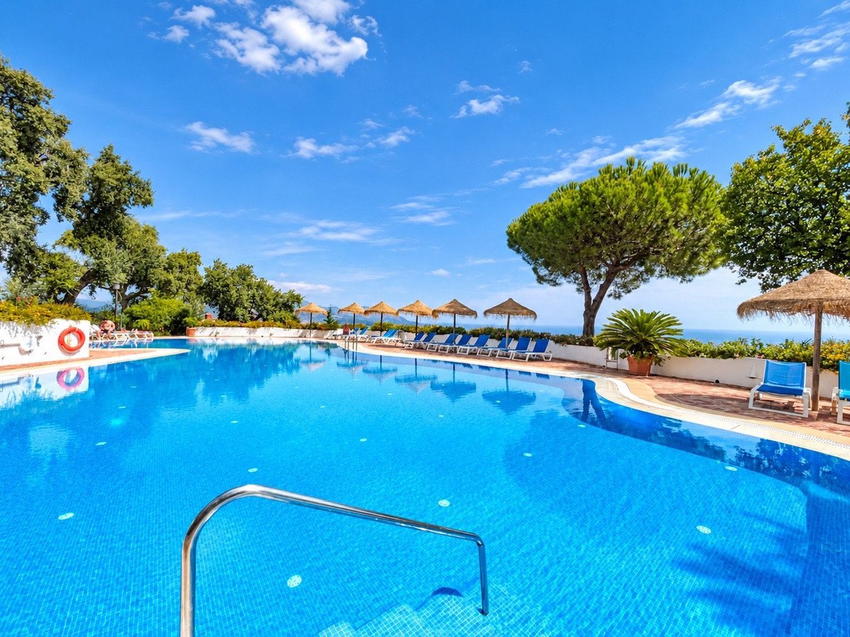 Large blue swimming pool with lounge chairs and umbrellas, overlooking the sea under a clear blue sky.