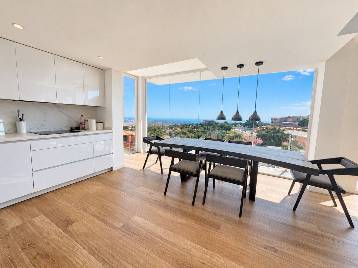 Modern white kitchen and dining area with large windows overlooking the sea and distant cityscape.